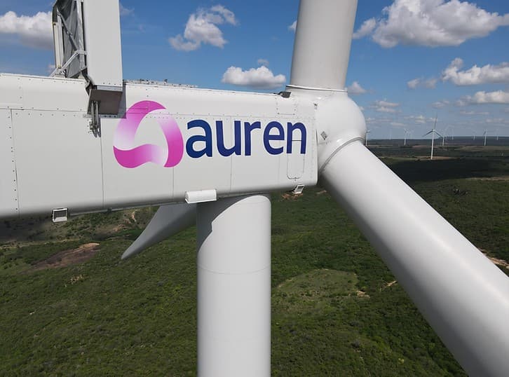 Turbina eólica com o logotipo da Auren Energia em destaque sobre fundo de mata e céu azul.