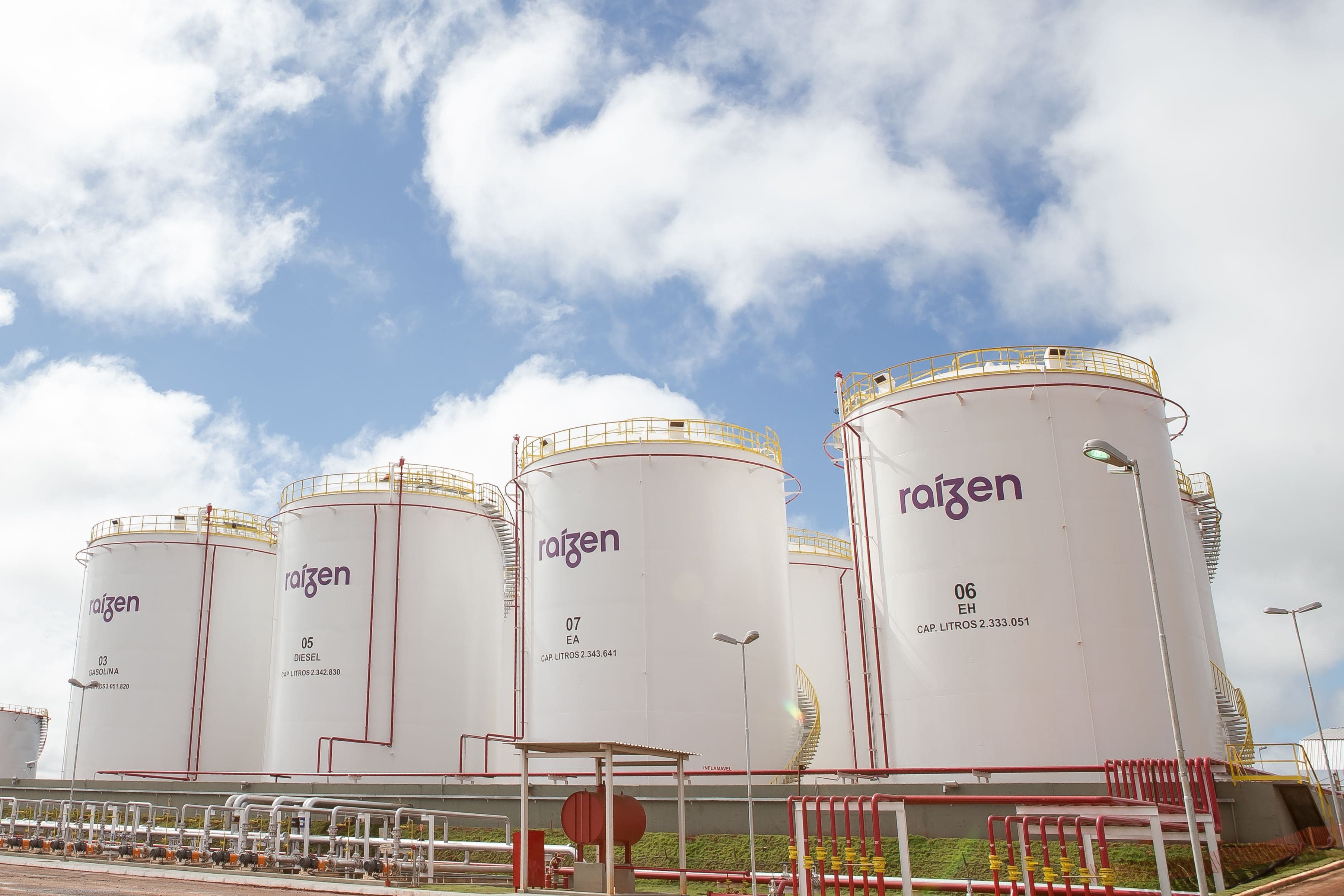Grandes tanques industriais brancos de armazenamento com o logotipo da Raízen sob um céu azul com nuvens.