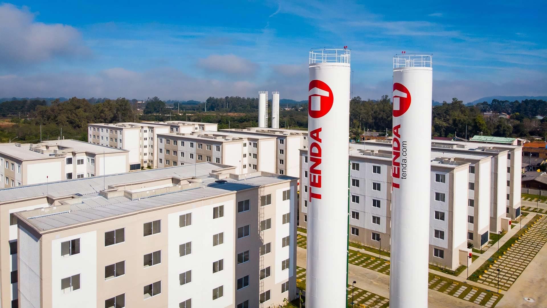 Vista de um condomínio residencial da Construtora Tenda, com prédios claros e duas torres de água ostentando o logotipo vermelho da empresa.