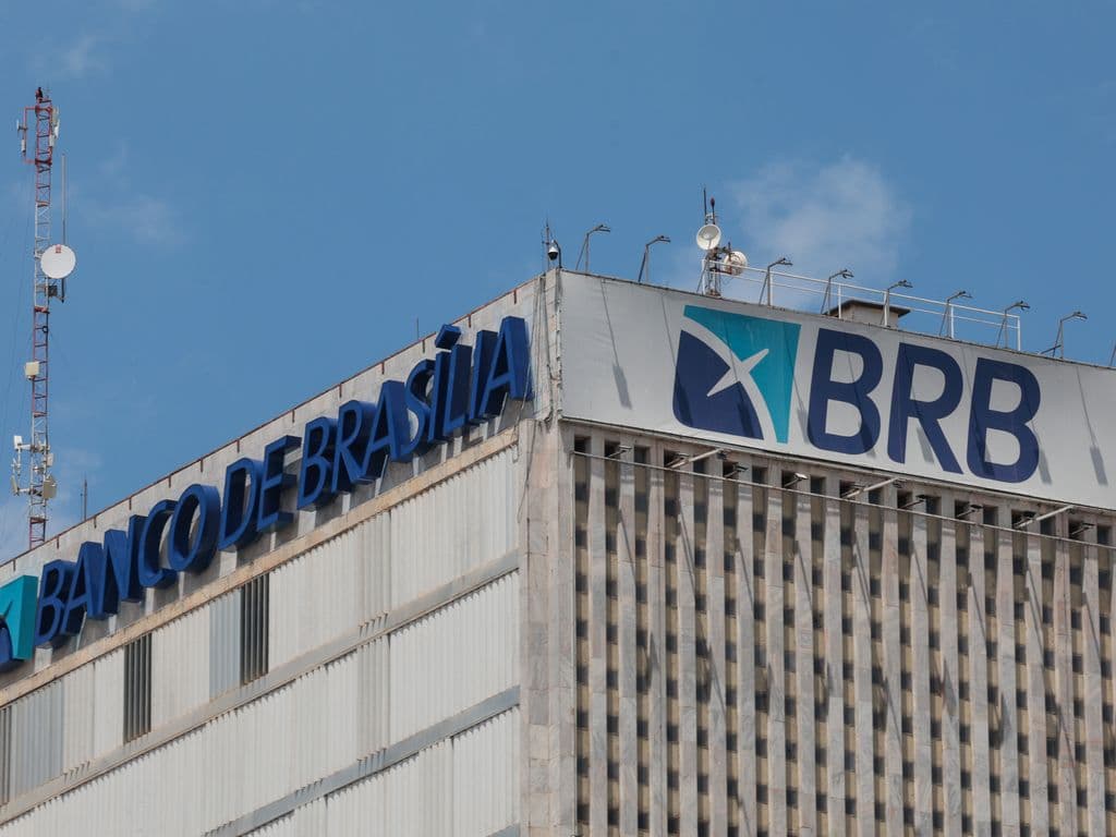 Fachada do topo do prédio do Banco de Brasília (BRB) com letreiros azuis indicando o nome e o logotipo da instituição sob céu claro.