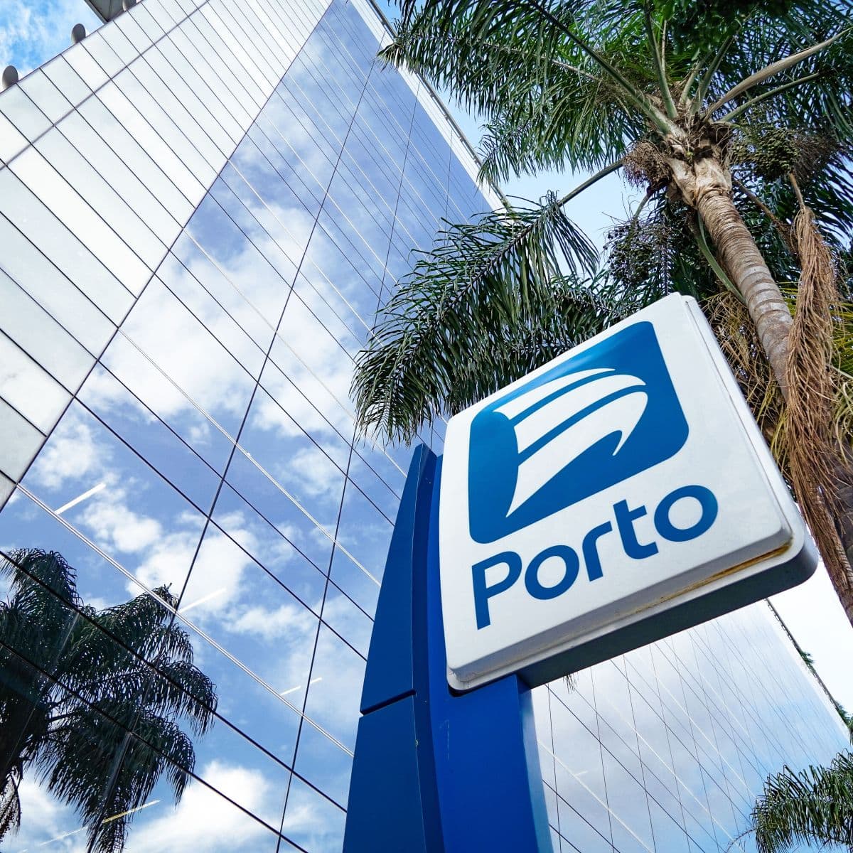 Placa com o logotipo da Porto em frente a um edifício corporativo espelhado e palmeiras.