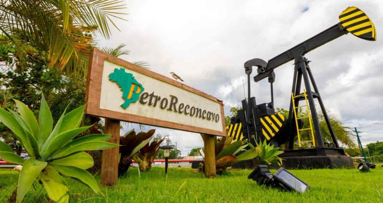 Placa de madeira com o logotipo da PetroReconcavo ao lado de um mini cavalo mecânico de extração de petróleo em um gramado verde sob céu nublado.